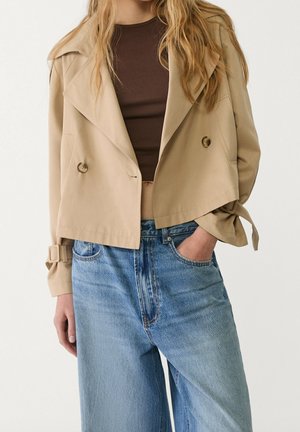 Vrouw draagt een beige korte trenchcoat over een bruin topje met lichtblauwe high-waisted jeans, één hand in de zak, staand tegen een witte achtergrond.
