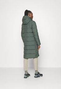 Groene pufferjas met een capuchon, tot op de knieën, met een doorgestikte design, gestroomlijnde silhouet, gecombineerd met beige broeken en zwarte schoenen.