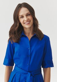 Blue button-up dress with short sleeves, collar, fitted waist, and tie detail. Smooth fabric with a solid color finish.