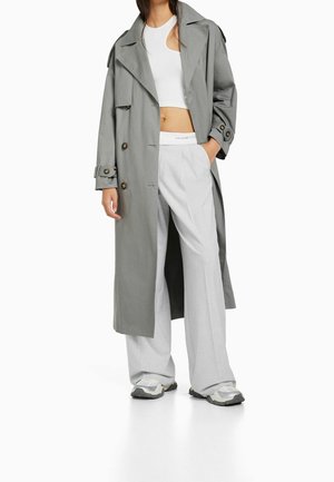 Frau trägt einen grauen langen Trenchcoat, ein weißes Crop-Top, weit geschnittene hellgraue Hose und weiße Sneakers, steht mit einer Hand in der Tasche.