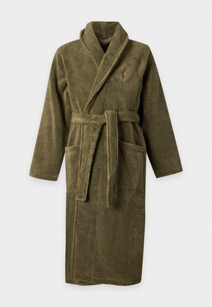 Homme debout pieds nus portant un long peignoir en peluche vert olive avec des poches et une ceinture sur fond blanc uni.