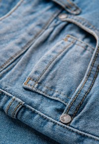 Tissu en denim bleu clair avec coutures visibles. Comprend une petite poche et un bouton en métal gravé "OH3BRE". Surface douce et texturée.