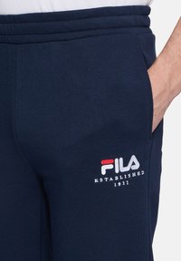 Granatowe dresy o miękkiej teksturze, z haftowanym logo FILA w czerwonym i białym kolorze oraz bocznymi kieszeniami na drobiazgi.