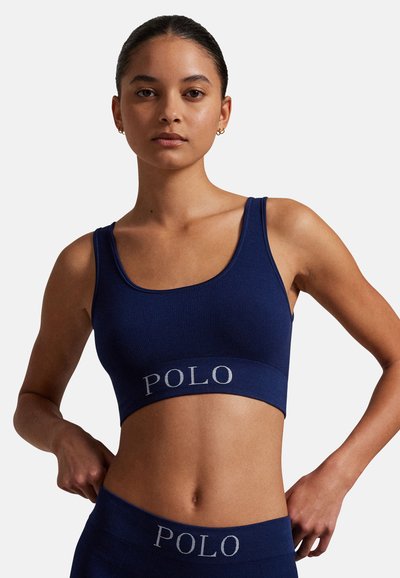 Tmavě modrá sportovní podprsenka s žebrovaným vzorem a širokou elastickou páskou s nápisem "POLO". Hladký materiál s kulatým výstřihem a nastavitelnými ramínky.