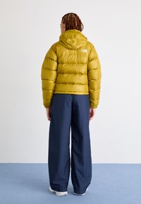 Rumena puhas jakna z elastičnimi manšetami in kapuco, oblečena čez ohlapne mornarsko modre hlače. Lahka tkanina s sijajnim zaključkom.