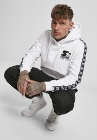 Hanorac alb cu logo negru și dungi laterale, asortat cu pantaloni negri și adidași albi. Materialul pare moale, cu detalii vizibile de cusătură.
