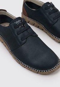 Zapatos de cuero azul oscuro con cordones elásticos, superficie texturizada, suela de goma beige y detalle de cuero marrón en el talón. Presente un detalle de costura.
