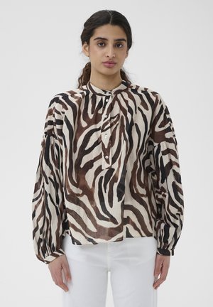 Femme portant un chemisier ample à manches longues avec des rayures zébrées marron et noires, boutonné au col, associé à un pantalon blanc.