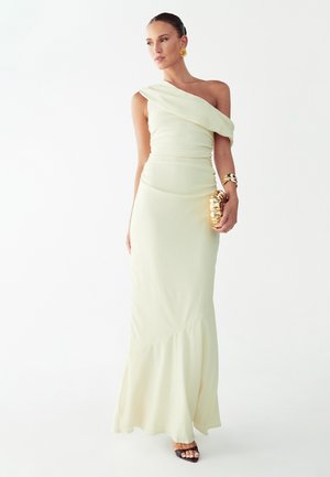 Vrouw in een crèmekleurige een-schouderjurk met een gouden clutch, geaccentueerd met gouden oorbellen en armband, staand tegen een witte achtergrond.