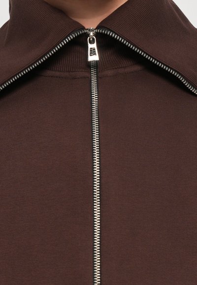 Braune Zip-Jacke mit hohem Kragen und silbernem Reißverschluss. Der Stoff ist glatt und gerippt, was das taillierte Design unterstreicht.