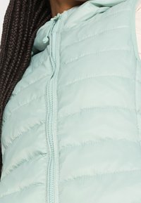 Light mint green puffer vest with a zip closure, hood, and diagonal stitching. Smooth texture, lightweight material, and no visible hardware.