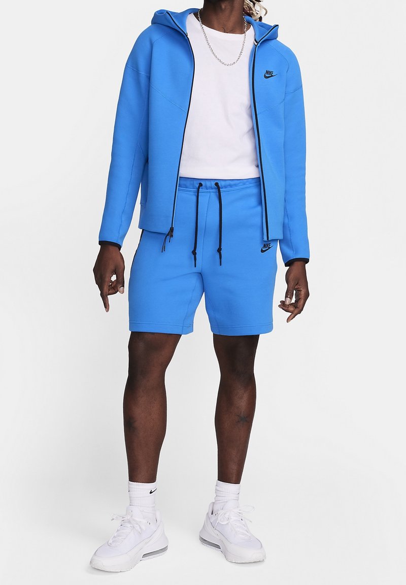 Person som bär en vit t-shirt, en blå Nike zip-up hoodie, matchande blå shorts, vita Nike strumpor och vita sneakers.