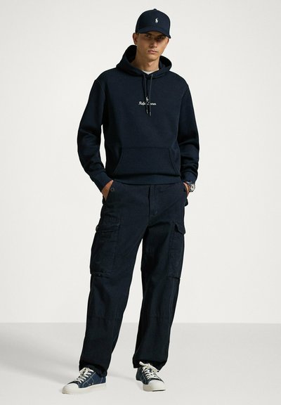 Felpa con cappuccio blu navy con logo, abbinata a pantaloni cargo scuri con tasche laterali. Completa il look con sneakers blu navy e un cappello coordinato.