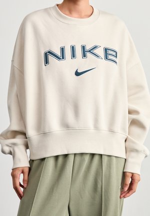 Osoba w oversize beżowej bluzie Nike z granatowym logo i oliwkowozielonych spodniach, stojąca na jednolitym tle.