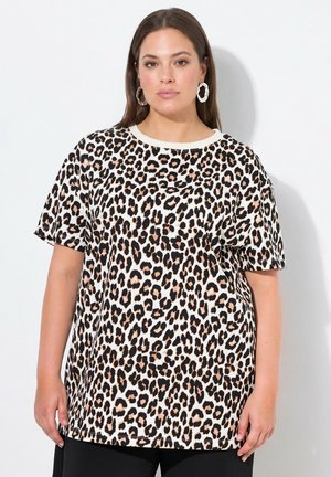 Vrouw met steil bruin haar draagt grote oorringen en een losse kortemouwen shirt met luipaardprint tegen een witte achtergrond.