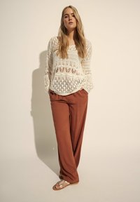 Jasnobeżowy sweter na szydełku z długimi rękawami, połączony z luźnymi spodniami w kolorze rdzy i jasnobrązowymi sandałami, stojący na neutralnym tle.