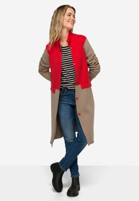 Cardigan lungo rosso e beige con una superficie testurizzata, dotato di bottoni lungo la parte anteriore. Indossato con una camicia a righe e jeans scuri.