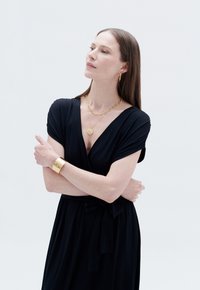 Abito nero avvolgente in morbido tessuto, scollatura a V, maniche corte, vita cinched con un fiocco, abbinato a gioielli e accessori dorati.