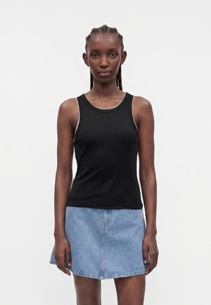 Young woman with braided hair wearing a black sleeveless top and light blue denim skirt, standing against a plain white background.