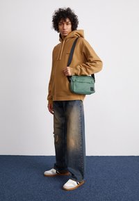 Grøn messenger-taske med sorte detaljer, bæres som crossbody. Brun hættetrøje, slidte blå vide jeans og hvide sneakers.