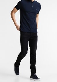 Polo navy de dos botones, con corte ajustado, combinado con jeans negros y zapatillas navy. La tela es suave, con un sutil detalle del logo en el pecho.