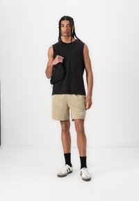 Sort ærmeløs skjorte, beige teksturerede mesh shorts, hvide og grå sneakers, sorte strømper og en lille sort taske. Enkel, sporty design.