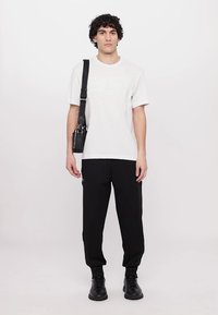 Biela bavlnená tričko s vyrazeným logom, čierne fleecové nohavice so zipsom na boku a čierna obuv. Čierna crossbody taška visí z ramena.