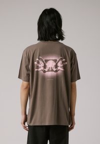 Brown cotton t-shirt featuring a graphic design of two pink hands in front of a faded image. Loose fit with a crew neckline.