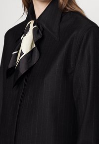 Svart ränder blåsa med en stor, volangkrage som pryds av en krämfärgad och svart mönstrad scarf som accent. Slät tygtextur.