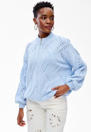 Kobieta w jasnoniebieskim, perforowanym swetrze na zamek i białych spodniach z kwiatowymi wycinankami, pozująca z jedną ręką w kieszeni.