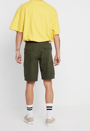 Żółta, oversize'owa koszulka zestawiona z ciemnozielonymi szortami cargo z bocznymi kieszeniami. Białe skarpetki w czarne paski i jasne buty.