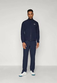 Navy zip-up jacket and matching pants in a soft fabric. Features a logo on the jacket. Model wears white sneakers with blue accents.