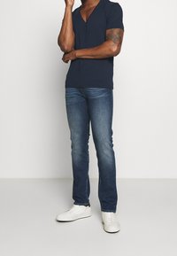 Uomo che indossa una maglietta blu navy a V, jeans blu e sneakers bianche, in piedi con un braccio incrociato e l'altra mano che tocca il viso.