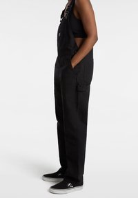 Schwarze Denim-Overalls mit verstellbaren Trägern, Seitentaschen und einem lockeren Schnitt, kombiniert mit schwarzen Slippern.