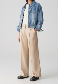 Veste en denim avec un lavage bleu, fermeture par boutons et poches à rabat, associée à un pantalon ample beige et des chaussures noires.