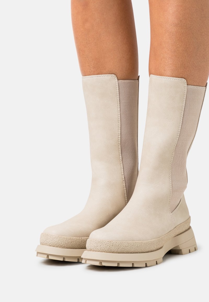 Paire de bottes beige mi-mollet avec semelles épaisses et panneaux latéraux élastiques texturés portées sur jambes nues sur un fond uni.