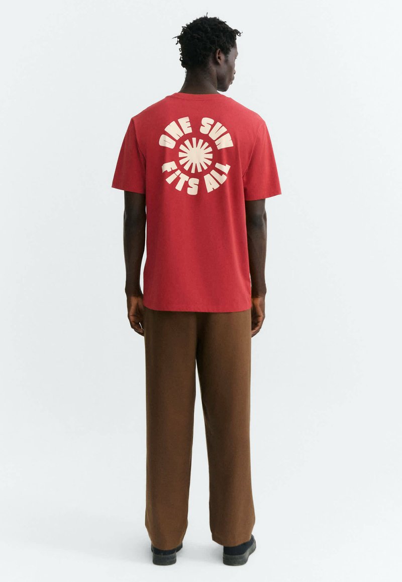 Un homme est debout de dos, portant un t-shirt rouge avec "ONE SUN FITS ALL" et un graphique de soleil dans le dos, un pantalon marron et des chaussures noires.