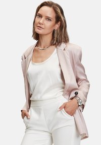 Lichtroze satijnen blazer over een witte satijnen top, gecombineerd met witte broeken. Inclusief zilveren sieraden en een horloge, wat zorgt voor een afgewerkte uitstraling.