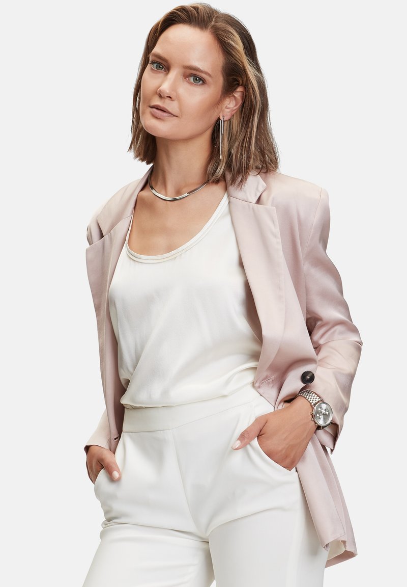 Lichtroze satijnen blazer over een witte satijnen top, gecombineerd met witte broeken. Inclusief zilveren sieraden en een horloge, wat zorgt voor een afgewerkte uitstraling.