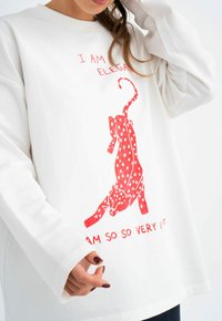 Witte sweatshirt met een rode afbeelding van een gestippelde kat, voorzien van tekst boven en onder in het rood. Het ontwerp heeft lange mouwen en een losse pasvorm.