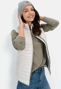 Leichtes, gestepptes beiges Weste mit hohem Kragen, graues Langarmshirt und graue gerippte Mütze mit einem Lederakzent. Denim-Jeans.