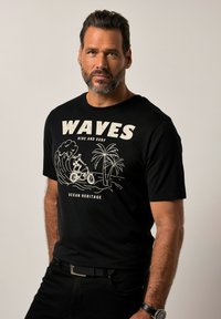 Zwarte katoenen t-shirt met een witte grafische afbeelding van een surfer, golven en palmbomen, met de tekst "WAVES RIDE AND SURF" erboven.