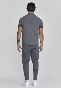 Camiseta atlética gris y pantalones de chándal con acentos negros, corte ajustado, combinados con zapatillas blancas; detalles de costura visibles y logotipo en los pantalones.
