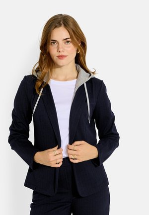 Joven mujer con traje de raya diplomática azul marino con capucha gris y camisa blanca, sosteniendo la parte delantera del blazer con ambas manos.