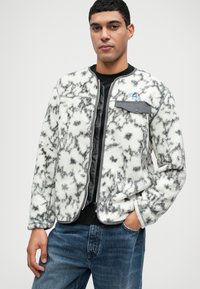 Fleecejakke i hvid og sort med et tekstureret, marmor mønster. Har en grå brystlomme og en lynlåslukning. Bæres over en sort skjorte.