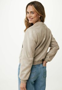 Jasno-beżowa kurtka bomberka z ekologicznej skóry, z prążkowanym kołnierzem i mankietami; charakteryzuje się gładką teksturą i dopasowanym krojem. Noszona z niebieskimi dżinsami.