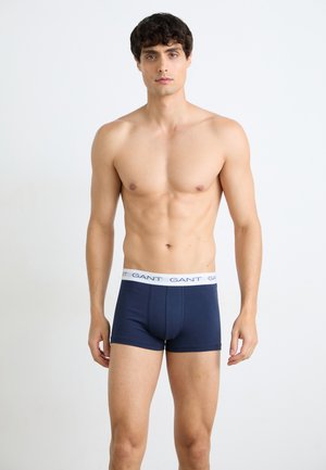 Man zonder hemd met kort krullend haar die donkerblauwe boxershorts draagt met een witte tailleband met het label "GANT", staat tegen een effen witte achtergrond.