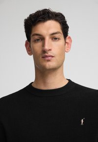 Suéter negro de punto acanalado con cuello redondo y un pequeño logo rosa en el lado izquierdo del pecho. Textura suave y diseño ajustado.