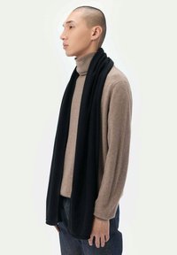 Svart scarf draperad runt en polotröja, tillverkad av mjukt material. Scarfen har en slät textur och en lång, rektangulär form.