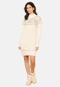 Abito maglione lavorato a maglia color crema con collo alto, motivi geometrici grigi e maniche lunghe. Abbinato a stivali alla caviglia beige foderati in pelliccia.